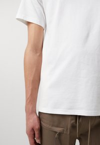 Hvid bomulds t-shirt med rund hals, parret med brune stofbukser med sidelommer og lynlåsdetaljer. Synlig hånd som holder om livet.