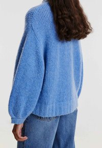 Personne avec de longs cheveux foncés portant un pull en tricot bleu clair ample et chunky, ainsi qu'un jean bleu à délavage moyen, dos à l'appareil photo.