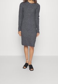 Robe grise en maille à manches longues avec une coupe ajustée, dotée d'un col rond et de détails à boutons sur l'épaule. Portée avec des chaussures à enfiler noires.