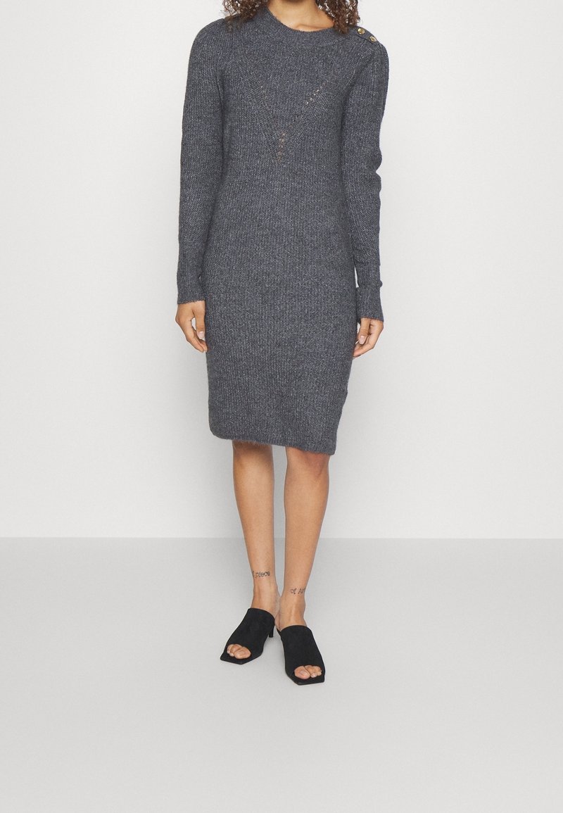 Robe grise en maille à manches longues avec une coupe ajustée, dotée d'un col rond et de détails à boutons sur l'épaule. Portée avec des chaussures à enfiler noires.