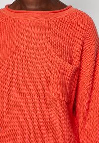 Pletený svetr v jasné korálové barvě se širokým výstřihem a krátkými rukávy. Na levé straně je jedna náprsní kapsa. Texturovaný vzor.