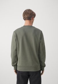 Uomo con capelli castani corti, che indossa una felpa verde oliva e pantaloni neri, girato di spalle su uno sfondo bianco uniforme.