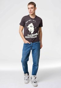 Black graphic t-shirt featuring a white portrait and text "Just ONE more thing!" paired with blue denim jeans and white high-top sneakers.