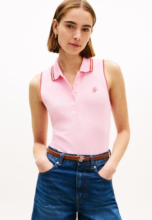 Mujer con una camisa polo sin mangas rosa con ribete rojo y jeans azules con un cinturón marrón, de pie con las manos en los bolsillos.