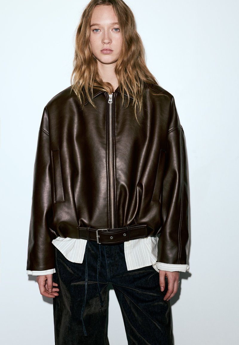 Veste en cuir marron avec fermeture éclair, coupe oversize, manches larges et ceinture à la taille, portée avec une chemise blanche à col en dessous.