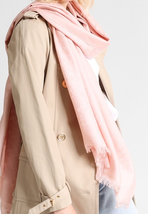 Écharpe rose pâle avec un motif subtil, bords effilochés, drapée sur un trench-coat beige, avec un bouton rond doré et un détail de manchette.