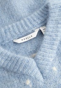 Miękki, jasnoniebieski sweter z dzianiny w białe groszki i białą metką marki "Noella" wewnątrz kołnierza.