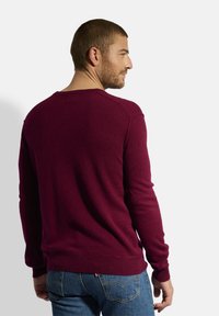 Bordeaux gestrickter Pullover mit langen Ärmeln und geripptem Saum, strukturierter Stoff und Rundhalsausschnitt. Über blauen Jeans getragen.