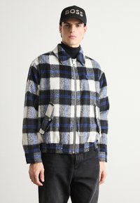 Plaid zip-up jacket in black, white, and blue with a fleece texture. Features side pockets and a collared design. Worn over a black turtleneck.