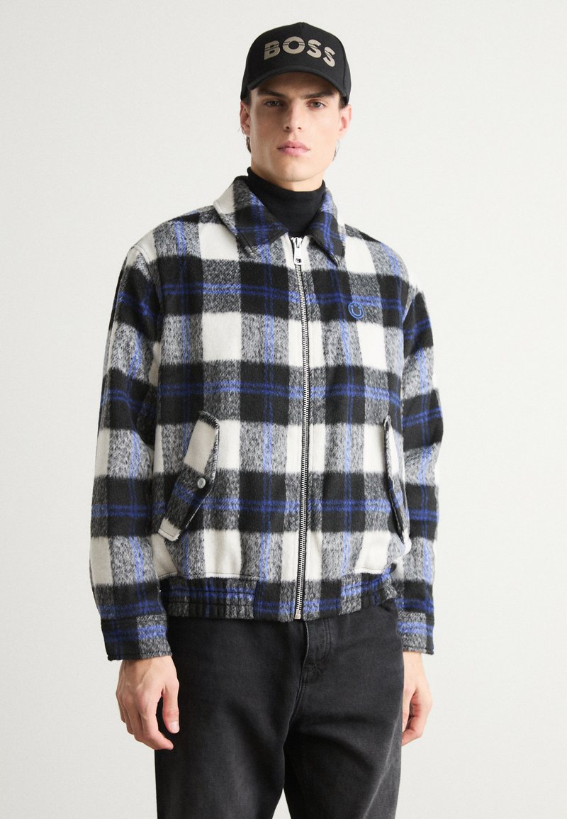Plaid zip-up jacket in black, white, and blue with a fleece texture. Features side pockets and a collared design. Worn over a black turtleneck.