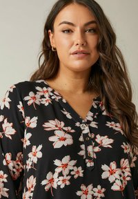 Blusa nera con motivo floreale bianco e rosso. Scollatura a V con dettagli di bottoni. Tessuto morbido, maniche lunghe e vestibilità rilassata.