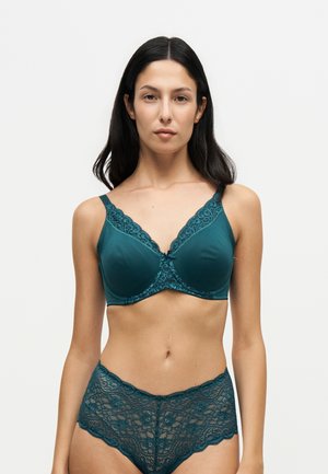 Femme aux longs cheveux foncés portant un soutien-gorge en dentelle turquoise et une culotte taille haute assortie en dentelle, debout devant un fond clair uni.