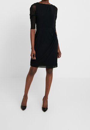 Frau, die ein knielanges schwarzes Kleid mit transparenten Dreiviertelärmeln und schwarzen spitzen High Heels trägt, steht auf einem weißen Boden.