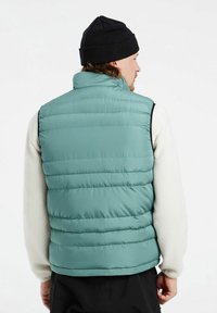 Kamizelka puchowa w kolorze teal, z pionowymi przeszyciami, wysokim kołnierzem i czarnymi akcentami. Nosi się ją na kremowym materiale polarowym.