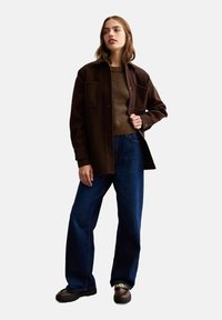 Giacca oversized marrone con chiusura a bottoni, due tasche frontali, indossata sopra un maglione marrone a trama e pantaloni larghi blu scuro. Scarpe marroni.