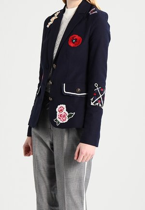 Marineblazer met bloemen- en ankerapplicaties, gedragen over een witte top en gecombineerd met grijze geruite broek.