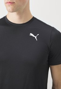 Muž v černém perforovaném sportovním tričku s bílým logem Puma na prsou, zobrazený od krku po horní část trupu na jednobarevném pozadí.