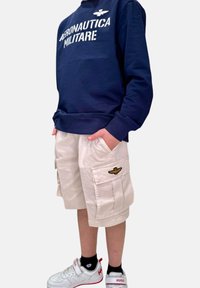 Felpa navy con testo bianco, pantaloni cargo beige con tasche laterali e emblema dorato, sneakers bianche e rosse, indossati con calzini neri.