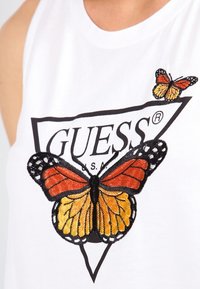Camiseta sin mangas blanca con un gran diseño de mariposa bordada en naranja y negro, con una mariposa más pequeña arriba y el logo "GUESS" abajo.