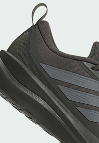 Gros plan d'une chaussure de sport noire avec une tige en tissu texturé, des bandes grises sur le côté, une semelle épaisse et rembourrée, ainsi qu'une languette au talon.