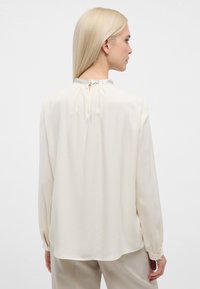 Flødefarvet bluse med lange ærmer, en nøglehulslukning bagpå og krave med sølvaccent. Blødt, flydende stof med glat tekstur.