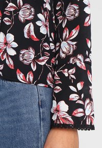 Blouse noire à motifs floraux blancs et rouges, avec une délicate bordure en dentelle sur la manche et un tissu doux et texturé.