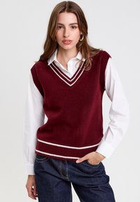 Gilet tricoté bordeaux avec un col en V, orné de rayures blanches le long du col et de l'ourlet, superposé sur une chemise blanche. Coupe décontractée avec une texture douce.