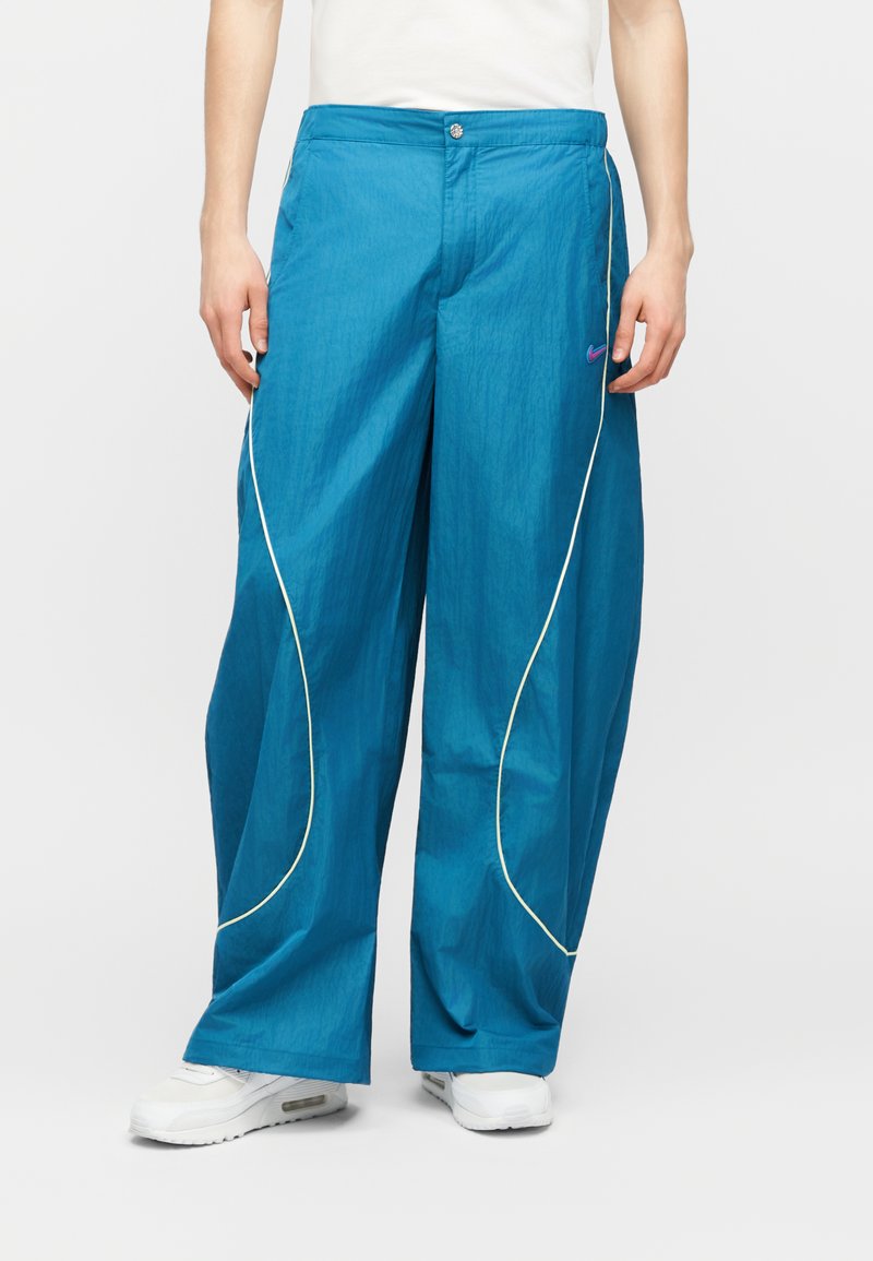 Pantaloni blu a gamba larga con piping bianco e un piccolo logo Nike viola, indossati con sneakers bianche e una maglietta bianca.