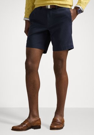 Shorts bleu marine en matériau léger, présentant une coupe droite et des ourlets retroussés. Inclut une ceinture en cuir brun et des mocassins bruns.