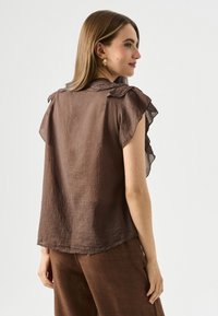 Blouse marron, légère, avec une texture, des manches à volant et un col à boutons. Le tissu a une finition chiffonnée, créant un look décontracté.