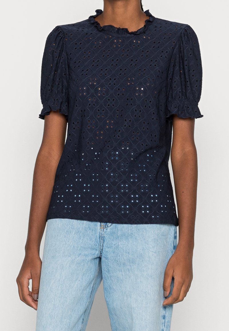 Blusa blu navy a maniche corte con colletto a volant; presenta un design a occhiello con motivi a diamante e aperture sottili. Vestibilità leggermente ampia.