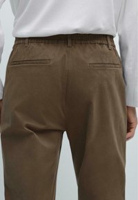 Bruine, elastische taille broek met een gladde textuur, voorzien van twee achterzakken en een relaxte pasvorm. Eenvoudig ontwerp zonder extra patronen.