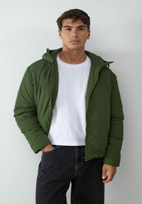 Giacca imbottita verde con zip frontale e cappuccio, indossata sopra una maglietta bianca, abbinata a pantaloni di jeans scuri. Texture liscia, design casual.