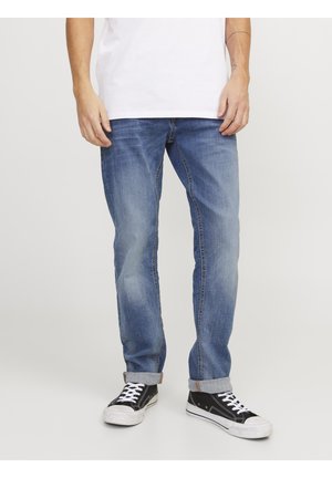 Person wearing blue rolled-up jeans, black and white sneakers, and a white shirt standing against a plain background.