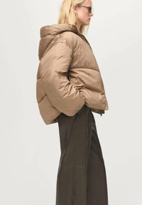 Giacca a vento marrone con cappuccio, caratterizzata da un design trapuntato e una chiusura frontale con zip. Il modello la indossa con pantaloni scuri a vita alta.