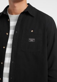 Juodas marškiniai su mygtukais ir krūtinės kišene. Savybė - Guess Jeans logotipo ženklas, dėvimi virš pilkos ir baltos dryžuotos T-marškiniais.
