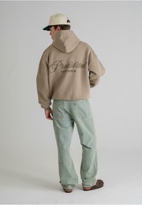 Beige oversized hettegenser med "Forbudt 10119 Berlin" trykk på ryggen, parret med lyseblå jeans og brune klomper på en nøytral bakgrunn.