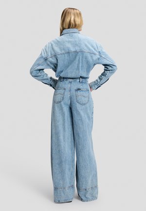 Person med langt blondt hår iført en løstsiddende lyseblå denimjakke og vidde jeans, står med hænderne på hoften med ryggen til.