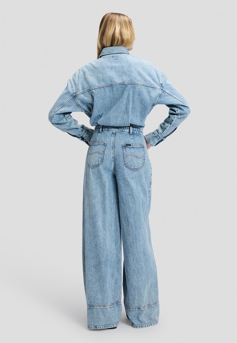 Personne aux longs cheveux blonds portant une veste en jean bleu clair ample et un jean à jambes larges, debout les mains sur les hanches, vue de dos.