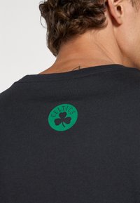 Sort bomulds T-shirt med en rund grøn lap med en kløver og teksten "CELTICS", placeret på bagsiden nær halsen.