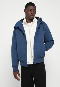 Man in een blauwe hoodie over een witte rits-top en zwarte broek, kijkend naar links met één hand in zijn zak van de jas.