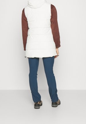 Personne portant une veste sans manches matelassée blanche, une chemise marron à manches longues, un pantalon bleu et des chaussures de randonnée, debout face à un mur blanc.