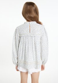 Lyseblå blomstret bluse med lange ærmer, med flæse-detaljer langs halsen og skuldrene, samt en nøglehulslukning bagpå. Hvide shorts nedenunder.