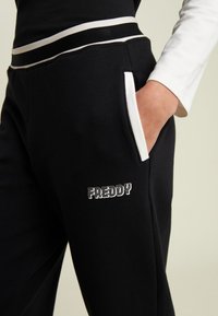 Pantalones de chándal negros con detalles blancos, un logo con la palabra "FREDDY" en el muslo, bolsillos laterales y una cintura elástica con rayas contrastantes.