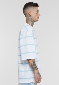 Tricou alb din bumbac cu dungi orizontale de culoare albastră deschisă, croială lejeră, umeri lăsați și decolteu rotund. Materialul pare moale.