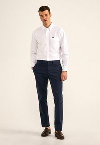 Camisa blanca de manga larga y botones con un bolsillo en el pecho, combinada con pantalones oscuros de corte ajustado color marino y mocasines marrones. Diseño simple y limpio.