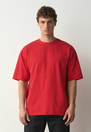 Junger Mann mit kurzen lockigen Haaren, der ein schlichtes rotes übergroßes T-Shirt und dunkle Hosen trägt, steht vor einem weißen Hintergrund.