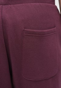 Ένδυμα από βυσσινί βαμβάκι με μια ραμμένη τσέπη, με λεία υφή και διακριτική γυαλάδα, τοποθετημένο σε φόντο απλής υφής.