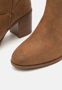 Bottine en daim marron avec un bout rond, une tige texturée et un talon bloc avec un motif de rayures horizontales. Fermeture éclair sur le côté.