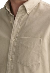 Beige kordflorsskjorta med knappar, ficka på vänster bröst och knäppta kragepunkter.